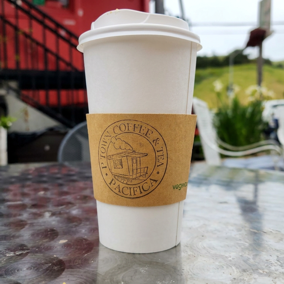Takeaway coffee cup labeled P-town Coffee & Tea Pacifica on an outdoor table overlooking a sunny green landscape.
